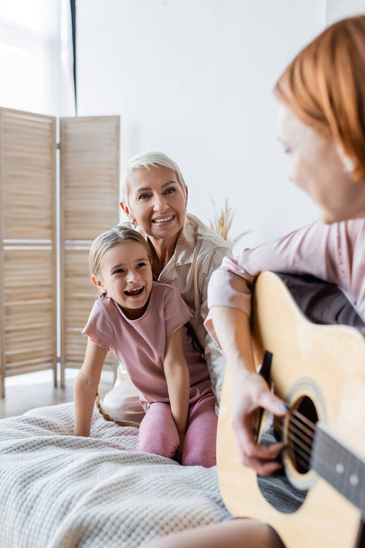 Lesbian woman and kid looking at blurred girlfriend playing acoustic guitar in bedroom  - 写真・画像