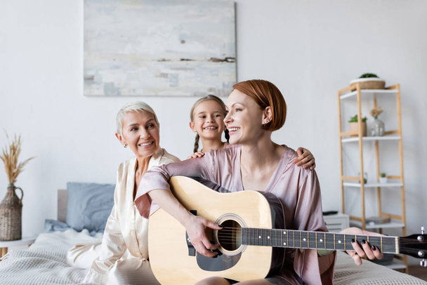 Χαμογελώντας γυναίκα και παιδί κοιτάζοντας φίλη παίζει ακουστική κιθάρα στο κρεβάτι  - Φωτογραφία, εικόνα