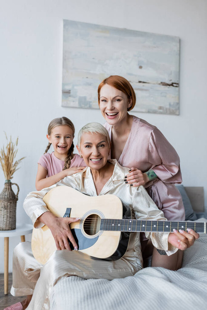Pozitív leszbikus pár akusztikus gitárral és lányával, akik otthon nézik a kamerát az ágyban  - Fotó, kép
