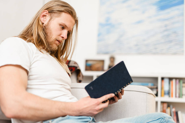 Homme blond barbu lisant son carnet tout en étant assis sur le canapé à la maison - Photo, image