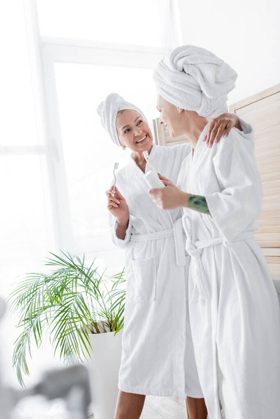 Smiling lesbian couple in bathrobes holding toothbrushes at home  - Фото, зображення