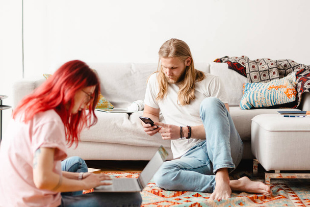 White man and woman using cellphone and laptop while sitting on floor at home - 写真・画像