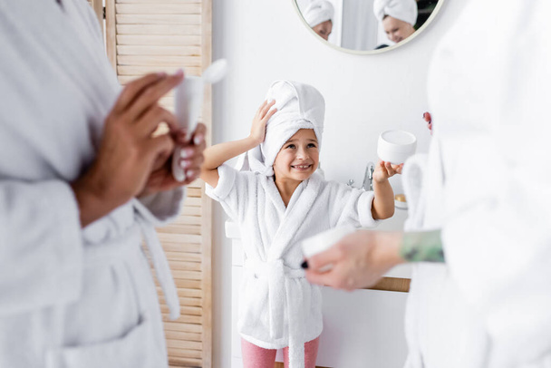 Pozytywne dziecko w ręczniku i szlafroku trzyma krem kosmetyczny w pobliżu matek w łazience  - Zdjęcie, obraz