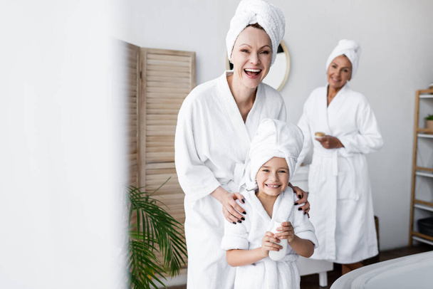 Cheerful woman hugging adopted daughter in bathrobe holding cream in bathroom  - Foto, imagen