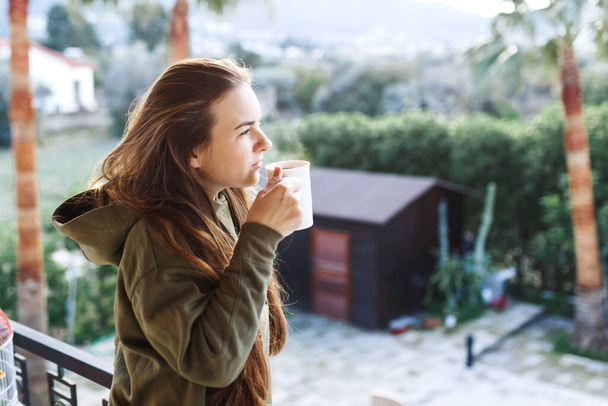 Portrét šťastné ženy, která pije kávu na balkonu.Uvolněte se, přemýšlejte a dívejte se jinam. Venku ve slunečním světle si užívá ranního čaje. V domě příroda měkká - Fotografie, Obrázek