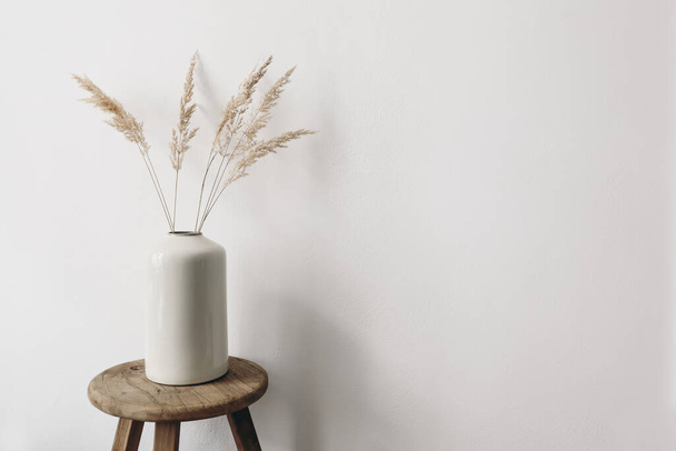 Modern summer, fall still life photo. Grey ceramic vase with dry festuca grass on old wooden stool. White wall background. Empty copy space. Elegant lifestyle decorative scene. Trendy interior decor. - Photo, image