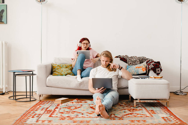 White man and woman using gadgets while sitting on couch and floor at home - 写真・画像
