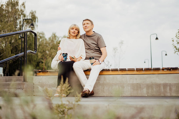 大人の幸せなカップルは、街の公園の屋外のベンチに座っているのが大好きです。ブロンドの白人男性と女性は一緒に時間を過ごし、コーヒーを飲む。外を歩く先輩妻と夫. - 写真・画像
