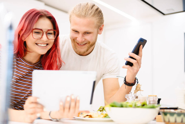 Young white couple using gadgets while having lunch in kitchen at home - Φωτογραφία, εικόνα