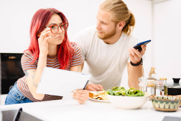 Young white couple using gadgets while having lunch in kitchen at home - Foto, Imagem