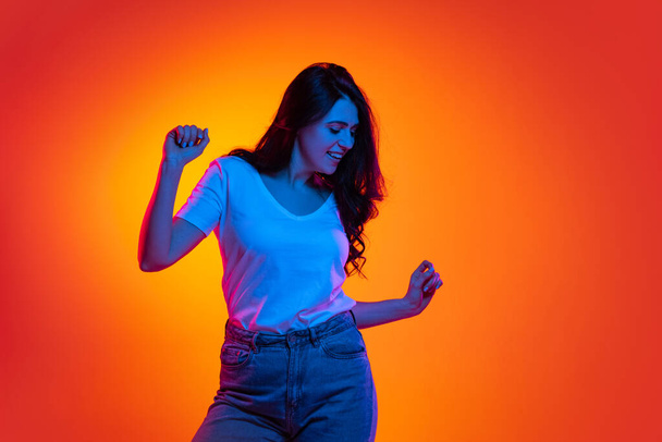 Excited young beautiful caucasian girl with long dark hair dancing isolated on orange background in neon light, filter. Concept of emotions, ads, sale - Fotografie, Obrázek