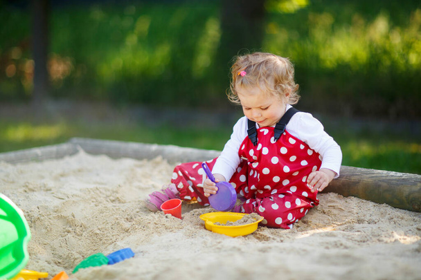 Roztomilé batole dívka hraje v písku na venkovním hřišti. Krásné dítě v červených gumových kalhotách baví na slunném teplém letním dni. Dítě s barevnými písečnými hračkami. Zdravé aktivní dítě venku hraje hry - Fotografie, Obrázek