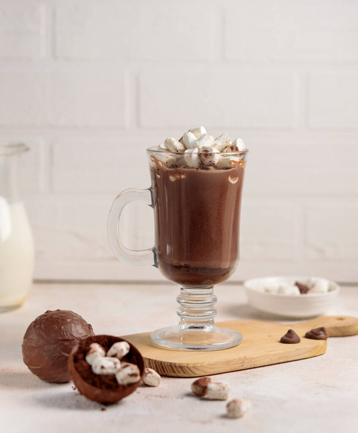 Cocoa in a tall glass mug and a chocolate bomb with a mini marshmallow for making a drink. Close-up, on a light brick background, selective focus - Zdjęcie, obraz