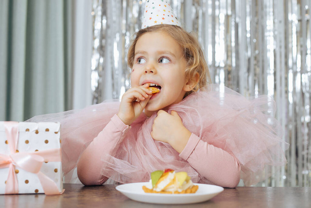 Little girl with short curly hair and stars on face in pink poofy dress and cap eating piece of fruit cake near gift box. - Foto, Imagen