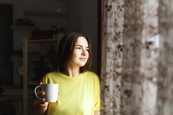 Giovane donna in piedi accanto alla finestra a casa e bere caffè. Scatto al coperto in stile casual. Mattina caffè donna routine quotidiana - Foto, immagini