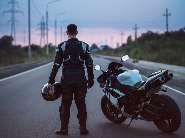 Un motociclista se para junto a una motocicleta en una carretera de asfalto y mira a la distancia. Puesta de sol de verano - Foto, imagen