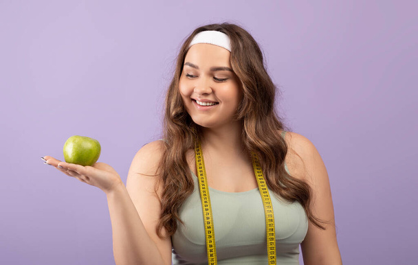 Veselý tisíciletý Evropan plus velikost samice ve sportovním oblečení s měřicí páskou drží zelené jablko na ruce - Fotografie, Obrázek