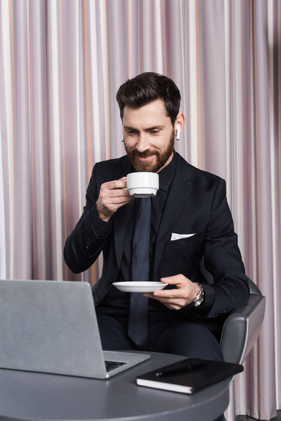 sonriente hombre barbudo en auriculares sosteniendo taza y mirando a la computadora portátil  - Foto, imagen