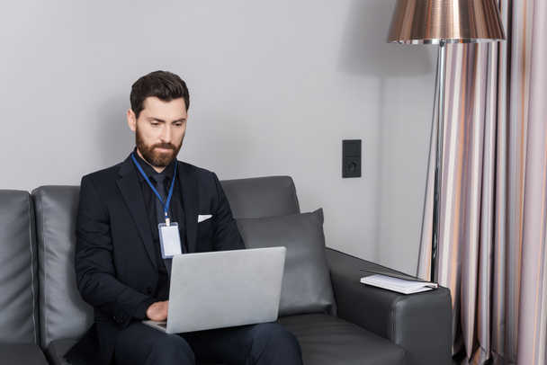 γενειοφόρος επιχειρηματίας με σήμα ταυτότητας χρησιμοποιώντας φορητό υπολογιστή, ενώ κάθεται σε δερμάτινο καναπέ στο δωμάτιο του ξενοδοχείου - Φωτογραφία, εικόνα