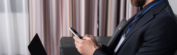 vista recortada de hombre de negocios con placa de identificación utilizando el teléfono inteligente cerca de la computadora portátil en la habitación del hotel, pancarta - Foto, Imagen