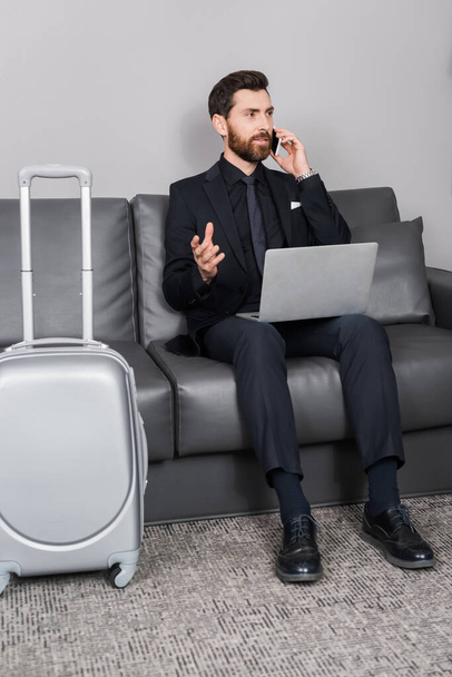 bearded businessman talking on smartphone near laptop and luggage in hotel room - Fotó, kép