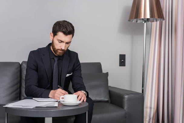 bearded businessman in suit writing on notebook near documents and cup of coffee in hotel room  - Фото, изображение