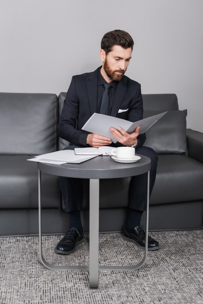 bearded businessman in suit looking at folder near cup of coffee in hotel room  - Φωτογραφία, εικόνα