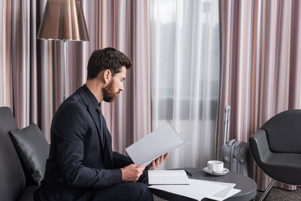 πλαϊνή άποψη του γενειοφόρου επιχειρηματία με κοστούμι κοιτάζοντας το φάκελο με χαρτιά κοντά φλιτζάνι καφέ στο δωμάτιο του ξενοδοχείου  - Φωτογραφία, εικόνα