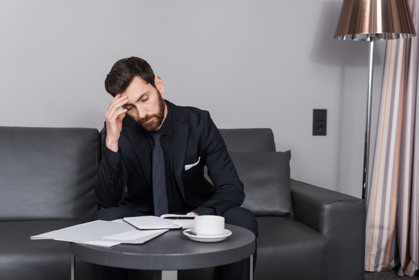 homem de negócios cansado em terno sentado no sofá perto de documentos e copo  - Foto, Imagem