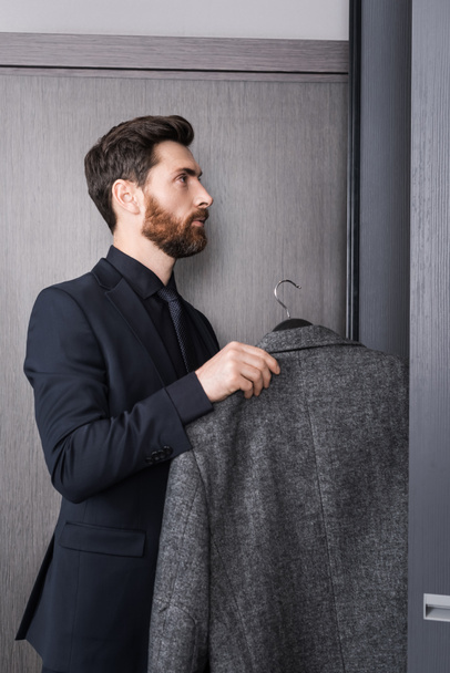 πλαϊνή άποψη του γενειοφόρου άνδρα στο κοστούμι κρατώντας κρεμάστρα με παλτό κοντά ντουλάπα στο δωμάτιο του ξενοδοχείου  - Φωτογραφία, εικόνα