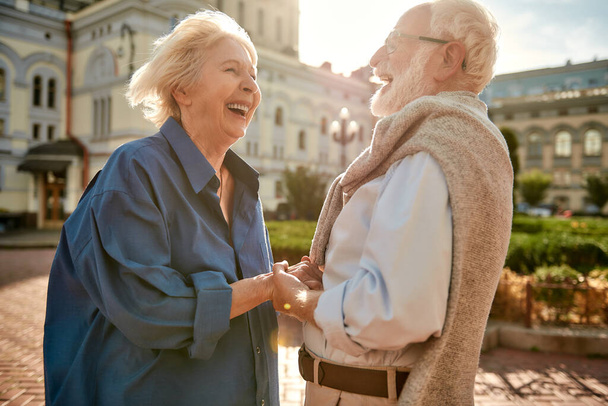 Happy and beautiful elderly couple holding hands and laughing while standing outdoors - Zdjęcie, obraz