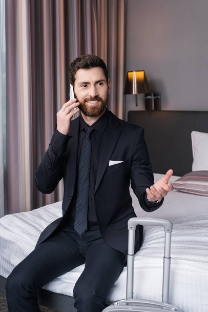 счастливый мужчина в костюме разговаривает по смартфону, сидя на кровати в гостиничном номере  - Фото, изображение