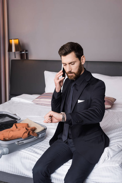 бородатый мужчина в костюме разговаривает по смартфону и проверяет время возле одежды в багаже на кровати - Фото, изображение