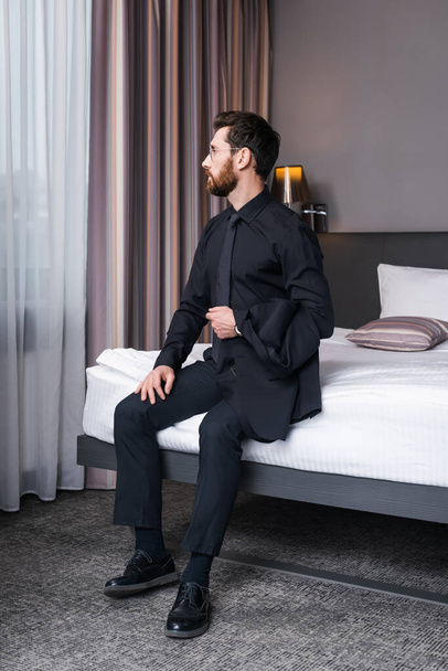 bearded man in eyeglasses and suit sitting on bed in hotel room  - Fotó, kép