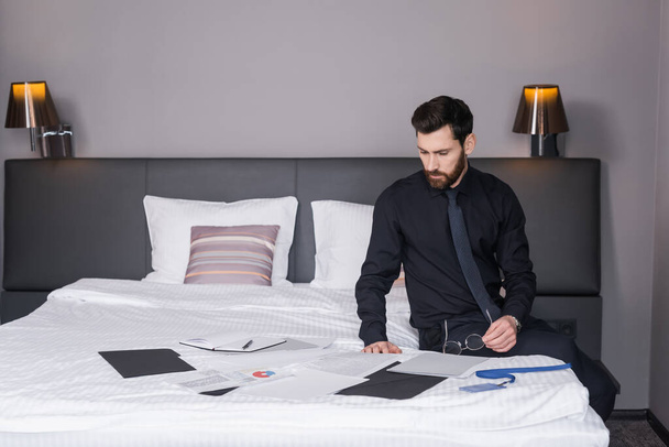 bearded businessman holding eyeglasses and looking at documents on hotel bed  - Foto, imagen