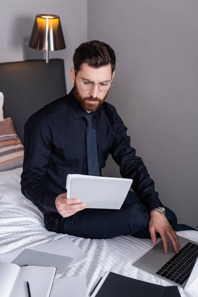 bearded man in suit and eyeglasses looking at folder and using laptop in hotel - Fotó, kép