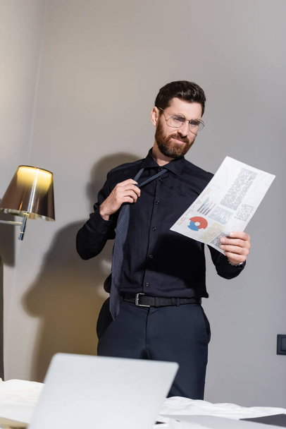 vousatý muž v brýlích upravující kravatu a dívající se na infografiku v hotelu - Fotografie, Obrázek