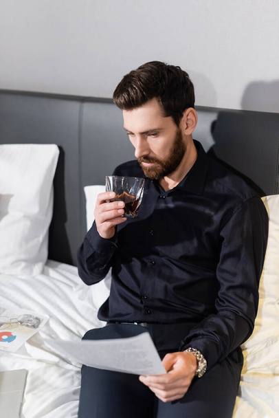 бородатый мужчина держит стакан виски и смотрит на размытый документ в отеле - Фото, изображение