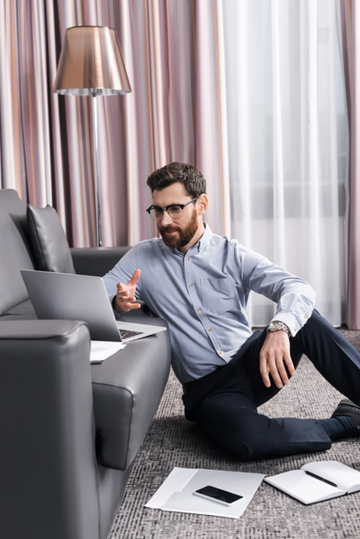 бородатый мужчина в очках сидит на ковре и разговаривает во время видеозвонка на ноутбуке  - Фото, изображение