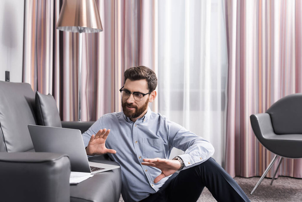 веселый мужчина в очках жестикулирует и разговаривает во время видеозвонка на ноутбуке  - Фото, изображение