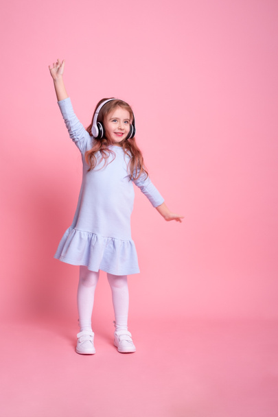 little girl in blue dress listening to music, dancing in big white headphones on pink background - Photo, Image