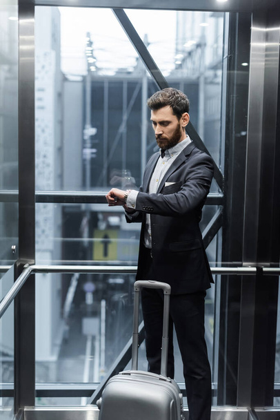 bearded man in suit checking time on wristwatch near luggage in modern elevator  - Foto, Imagem