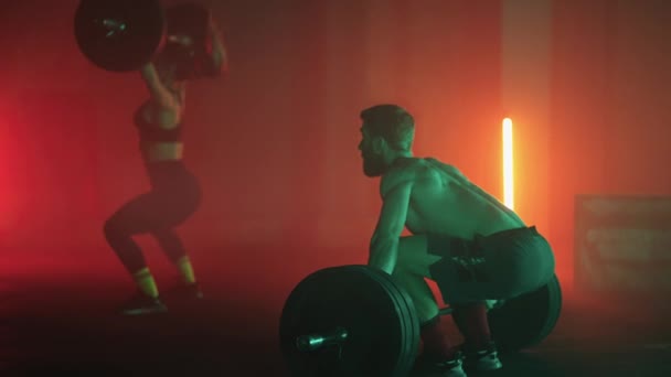 A man and a woman in a colored neon red and blue light in slow motion lift heavy barbells over their heads. Strength and power training with heavy weight. The concept of athletic and strong couples - Footage, Video