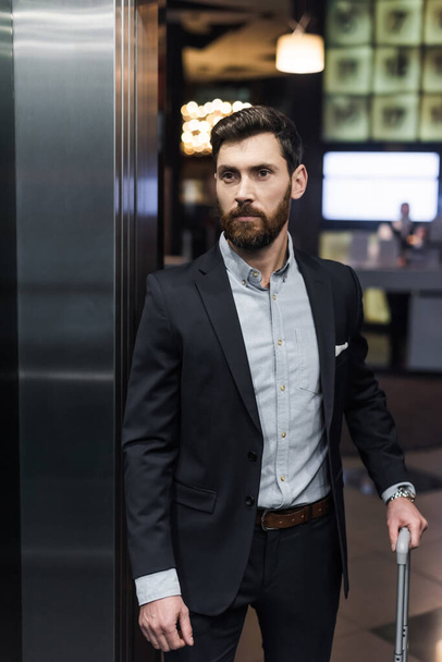 bearded businessman in suit holding luggage handle in modern elevator  - Valokuva, kuva