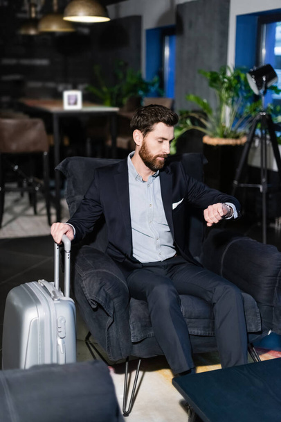 hombre barbudo en traje con asa de equipaje durante el registro de tiempo en el vestíbulo del hotel  - Foto, Imagen
