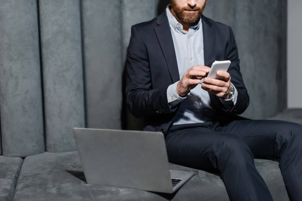 cropped view of bearded man in formal wear using smartphone near laptop in hotel lobby  - Fotoğraf, Görsel