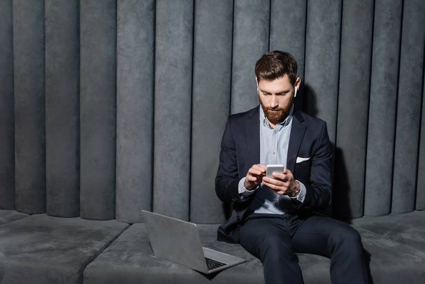 bearded man in earphones using smartphone near laptop in hotel lobby  - Zdjęcie, obraz