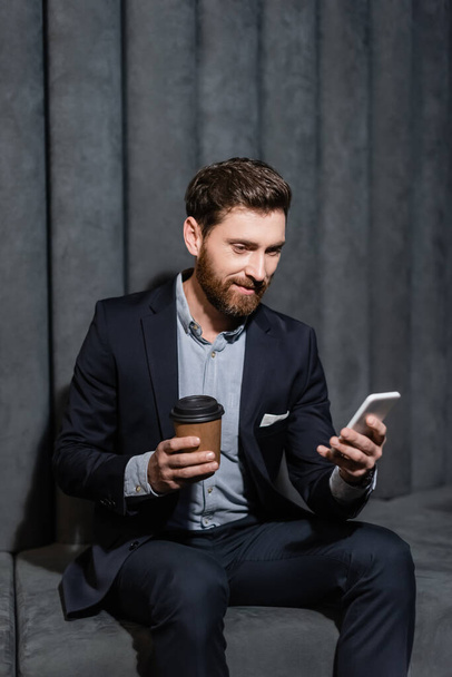 улыбающийся человек в костюме с помощью смартфона и с бумажной чашкой в холле отеля - Фото, изображение