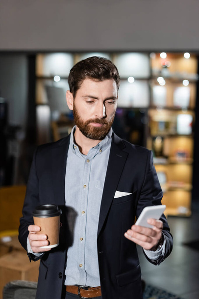 bearded man in suit holding smartphone and paper cup in hotel foyer - Foto, imagen