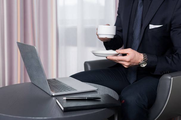 Modern otel odasındaki dizüstü bilgisayarın yanında takım elbiseli bir adamın elinde fincan ve çay tabağı vardı. - Fotoğraf, Görsel